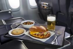Artisanal pretzel roll filled with tangy barbecue chicken and coleslaw paired with a tri-color quinoa side salad and beer, sitting on an airplane tray table.